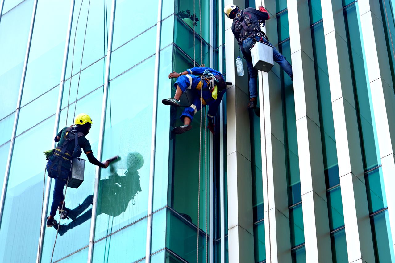 services-03 Three window cleaners suspended on ropes clean a modern glass building facade during the day.