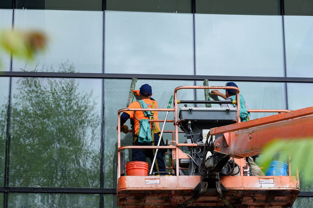 services-02 Two workers cleaning windows on a glass building facade using a lift platform, enhancing building aesthetics.